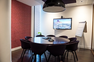 Kuva pienestä neuvotteluhuoneesta missä keskellä pöytä ja tuolit ympärillä sekä videoneuvottelulaite seinällä olevan näytön päällä.